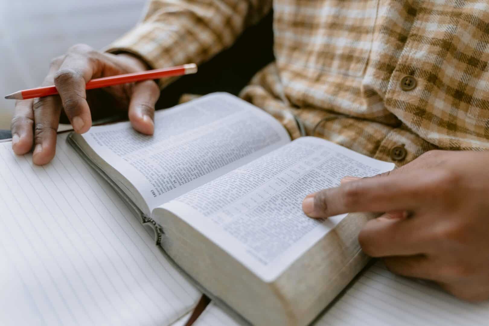 Hands holding an open Bible with a red pencil over a notebook